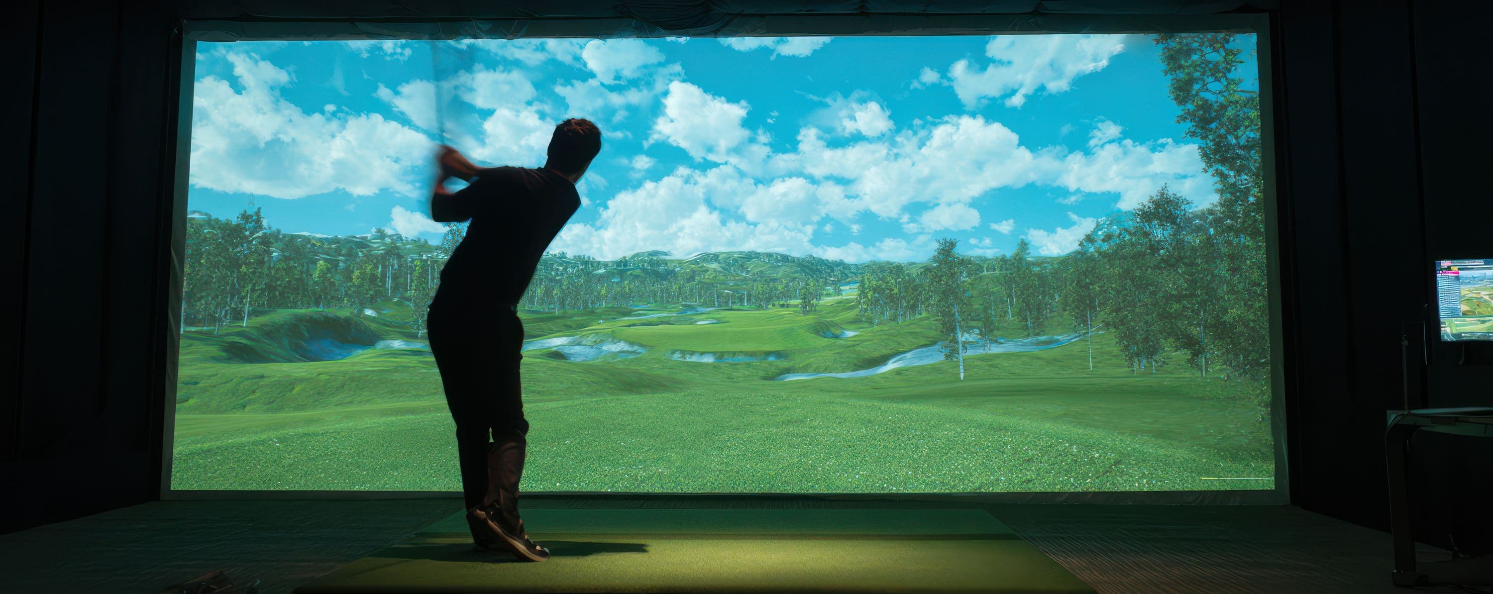A player mid-swing in a darkened simulator bay, silhouetted against the lit course on screen